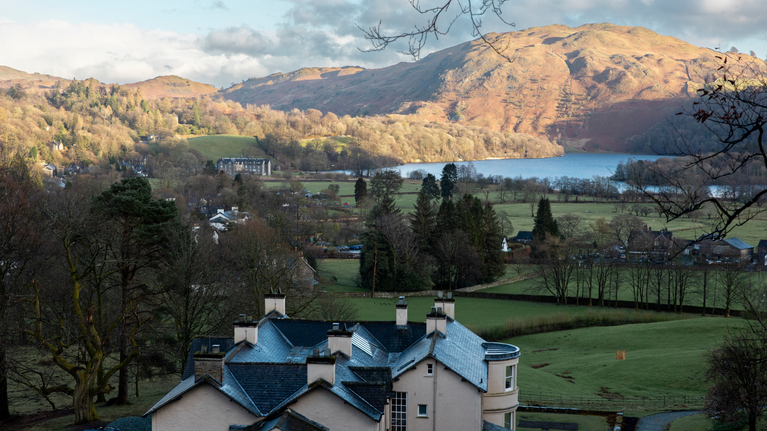 A historic house with slate roofs and chimneys surrounded by lush greenery, with a lake and sunlit hills in the background. A tranquil springtime woodland landscape.